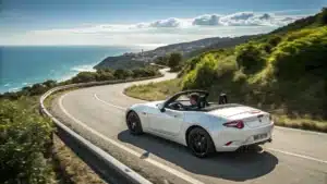 Une voiture décapotable stylisée (Mazda MX-5 par exemple) roulant sur une route côtière sinueuse avec un ciel dégagé et une mer scintillante à l’horizon.