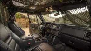 Int&eacute;rieur du Hummer H1 montr&eacute; en gros plan avec des touches de cuir et des &eacute;quipements technologiques, entour&eacute; d&rsquo;une ambiance militaire (filets, camouflage, terrain accident&eacute;).